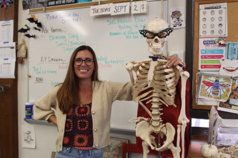 Art teacher Jillian Rodgers poses with her classroom skeleton, showcasing her knitted sweater with swirls of orange and maroon, preparing for spooky season.
