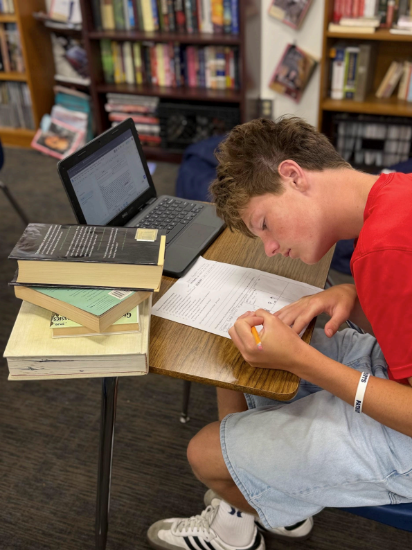 A student is seated in his desk working on a paper that appears to be homework, he is surrounded by a stack of books to represent the
constant studying from textbooks that is being required in this time of year, He also has his chromebook open to an assignment with tons of
writing to further the demonstration of work as he is surrounded by work in different forms piling on his desk and yet he still has to focus on each one in an aim for success.